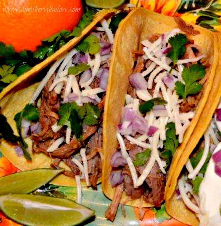 Two Cuban Street Tacos on a plate with an orange and 2 lime wedges. These tacos have orange and lime zest infused as the pork cooks along with Cuban inspired spices. The meat is in a tortilla, then shredded monterey jack cheese, diced red onion and cilantro.